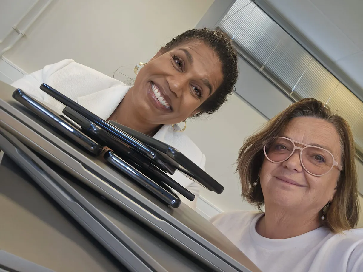 Twee vrouwen poseren met stapel laptops en telefoons.