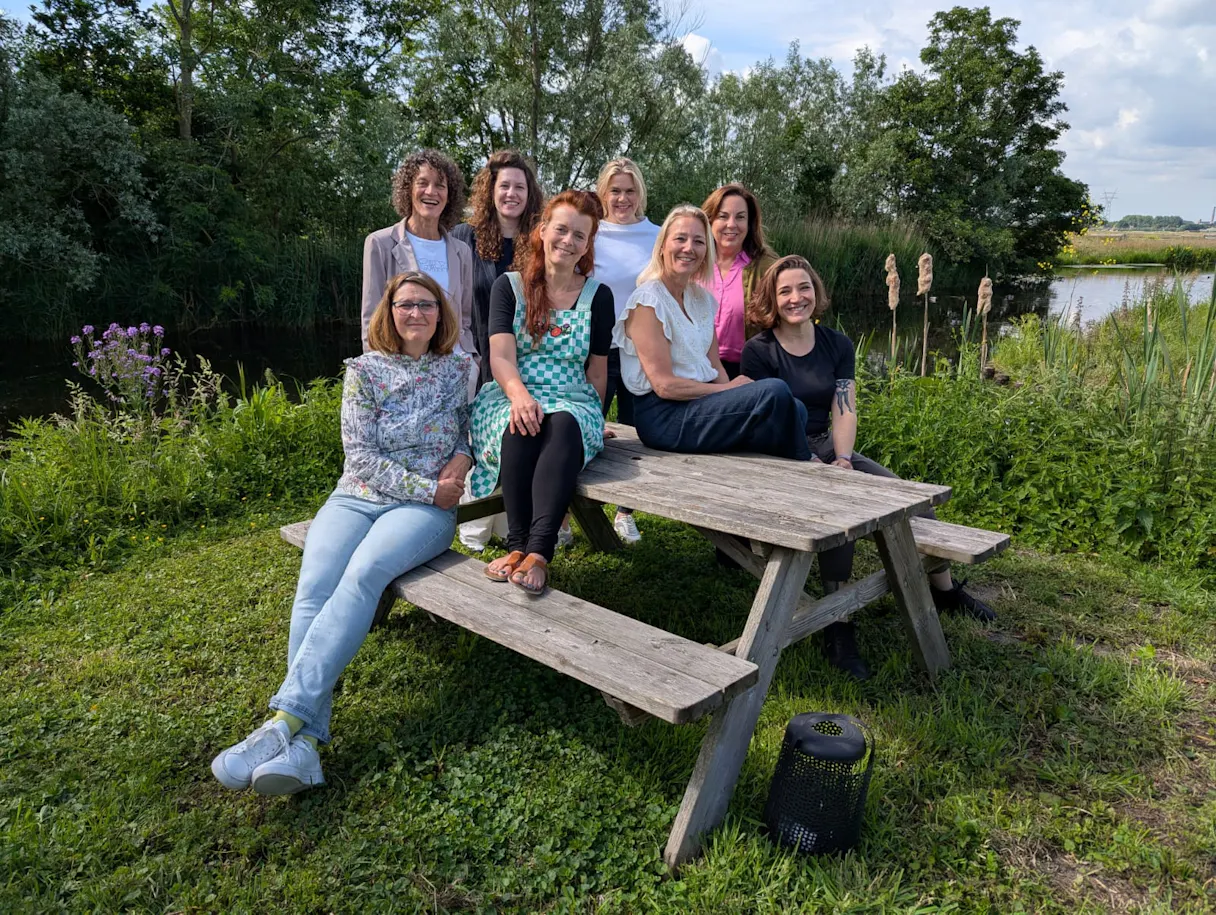 Groepsfoto van acht vrouwen buiten bij een vijver.