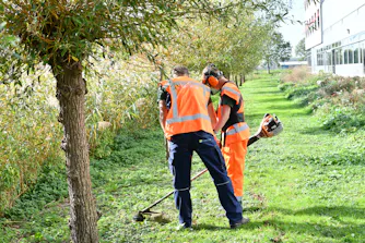 Twee groenmedewerkers maaien gras langs een gebouw.