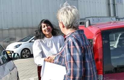 Vrouw schudt hand van man bij auto, lachend.