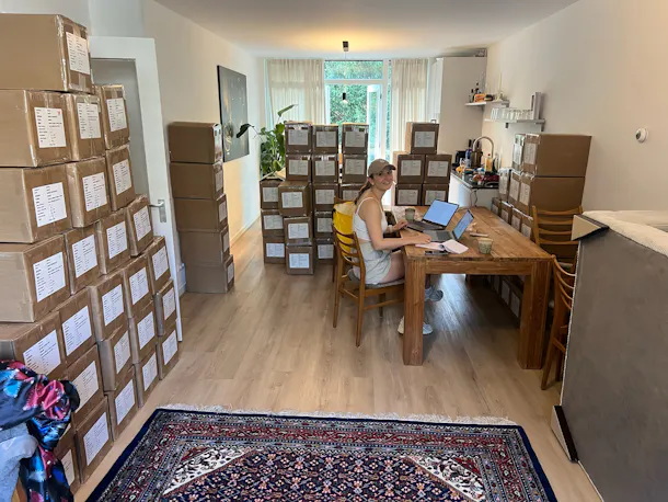 Vrouw achter laptop, omringd door dozen in kamer.