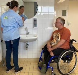 Vrouw helpt man in rolstoel bij wasbak.